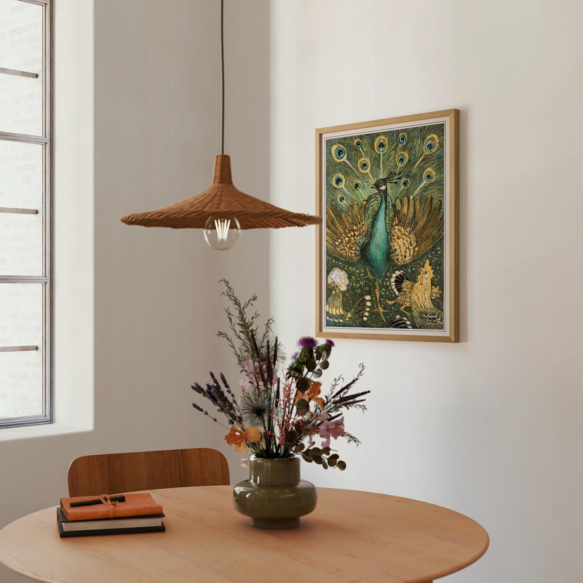 a wooden table with a vase of colorful flowers, a stack of books, and a hanging light fixture. The table is positioned next to a window, and a framed painting of a peacock is displayed on the wall above it.