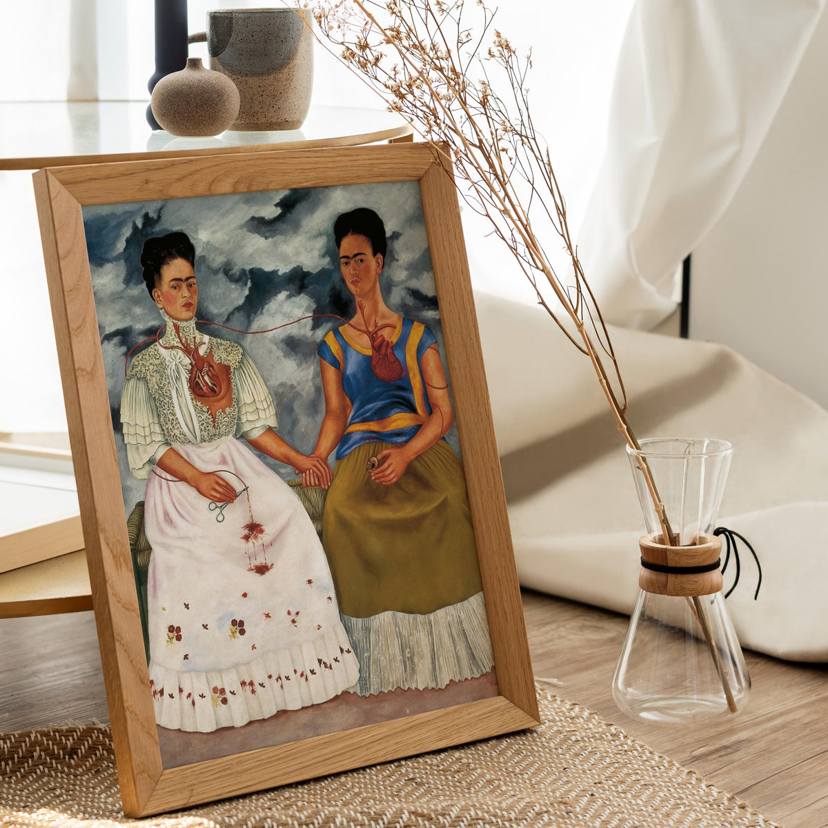 a framed painting of two women, one wearing a white dress and the other in a yellow dress, sitting together and holding hands. The painting is displayed on a wooden surface, with a vase of dried flowers and a glass vase with a wooden handle placed nearby.