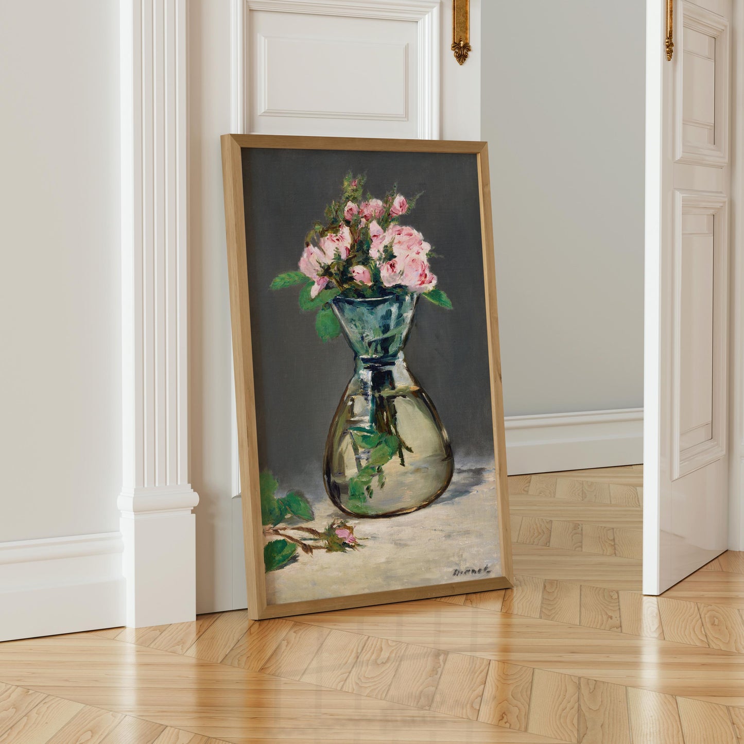 A framed painting of a vase filled with pink roses is leaning against a white wall in a room with a wooden floor.