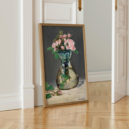A framed painting of a vase filled with pink roses is leaning against a white wall in a room with a wooden floor.